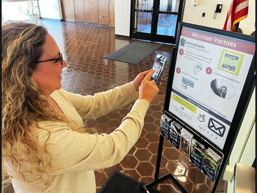 A woman with long blond hair, glasses and wearing a white shirt uses a mobile device to scan a QR code on a poster hanging outside a receptionist's window